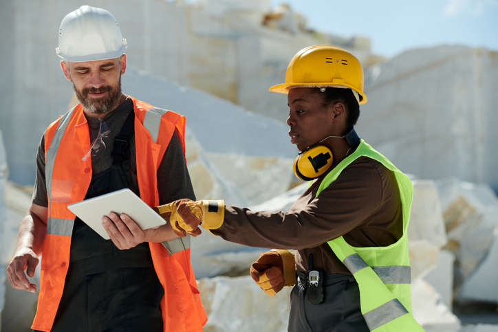 Young female engineer pointing at screen of tablet held by foreman in safety helmet and workwear during work on quarry